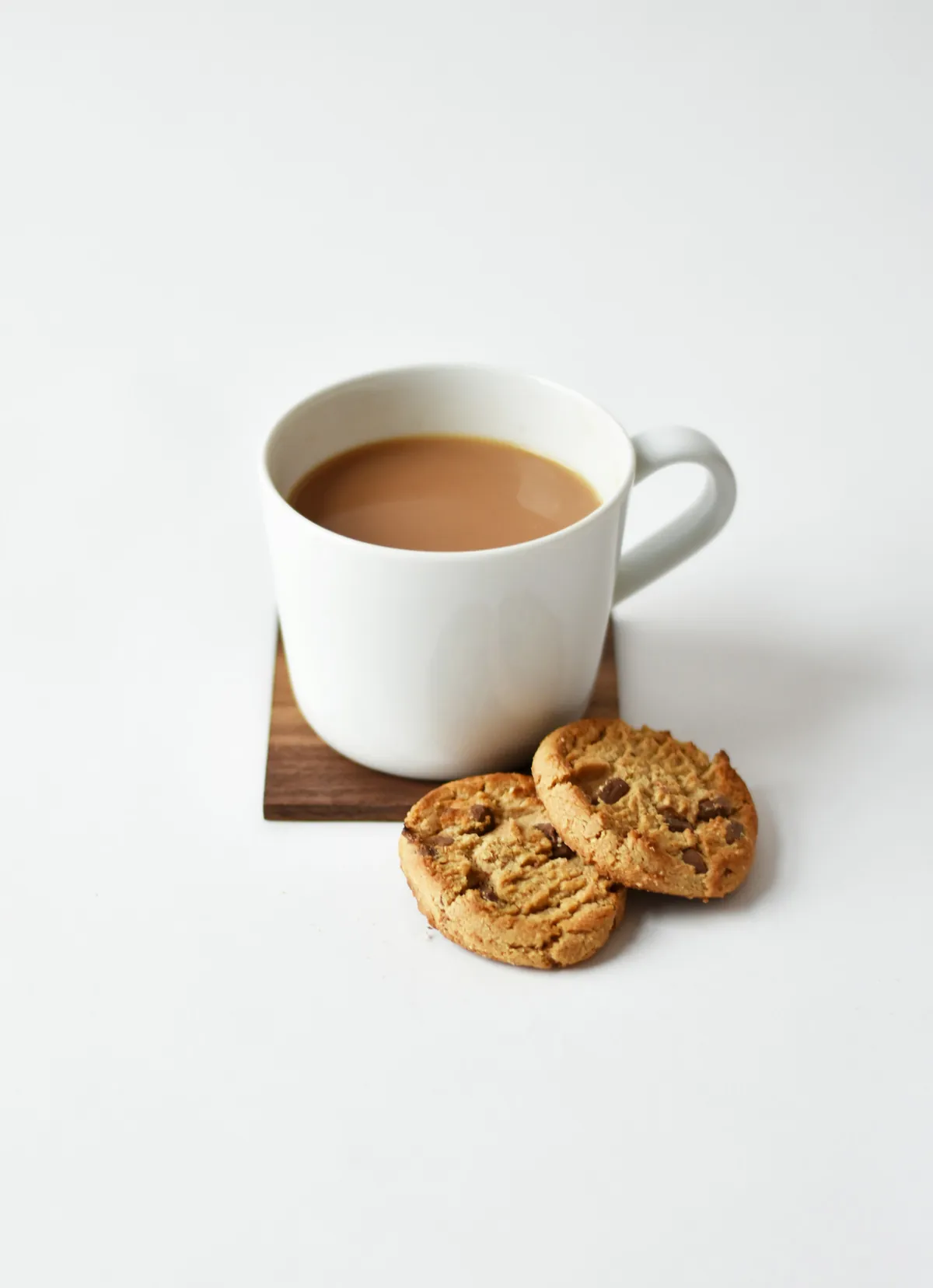 White cup of tea on a wooden coaster next to two cookies on a white background