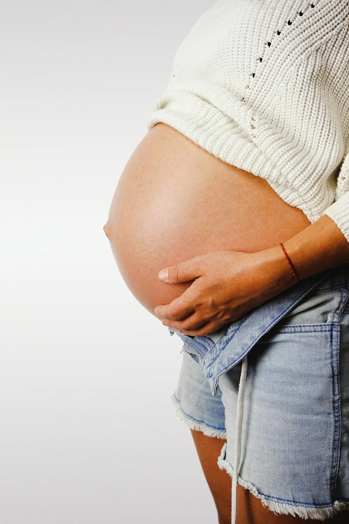 Pregnant woman in a white sweater and denim shorts holding her belly